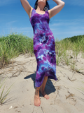 A woman wearing a sleeveless tie-dye maxi dress in purple, blue, and white stands on a sandy beach with dune grass and blue sky in the background.