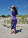 A woman stands on a sandy beach with dune grass and a blue sky behind her, wearing a sleeveless midi dress with a purple and blue tie-dye galaxy pattern.
