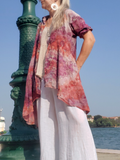 Person wearing a colorful shirt and white pants standing near a decorative green column with water and sky in the background.