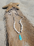 A beaded necklace with white pearl-like beads and a turquoise teardrop pendant, laid on a brown feather with a starfish on a sandy surface.