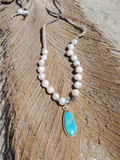 A beaded necklace with white freshwater pearls, a small silver-toned spacer bead, and a teardrop-shaped turquoise pendant set in silver, displayed on a textured brown palm leaf.