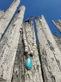A beaded necklace with white pearl-like beads and a turquoise teardrop pendant, draped over weathered wooden posts against a blue sky.