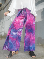 Person wearing tie-dye pants with a white top against a gray wall.