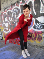 A cute and fun pose; Kelsie is pushing her upper body towards the camera, somewhat bent down. Her thumbs are sticking out the holes on the long sleeves,  arms up front of her, fists close to her face, the right front of the cardigan is  blowing in the wind.