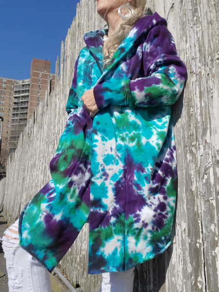 A very oversized big and extra long 100% cotton hoodie in hand dyed purple-green-jade-a bit of black. Open front, side pockets, rounded and unfinished raw hem, wide ribbed cuffs, hood. I'm leaning against a fence, holding the front together with one hand, the other one is in my pocket.