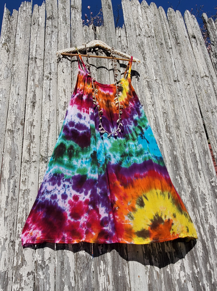 a very colorful & bright tie dye one sizebeach dress; parachute shape with adjustable straps. Rainbow Galaxy tie dye; jewllow-oj-pink-purple-green-jade-a bit of black. Very lightweight and breeze woven rayon fabric. Displayed on a hanger with one of my long shell & hemp necklaces, photographed on the fence.